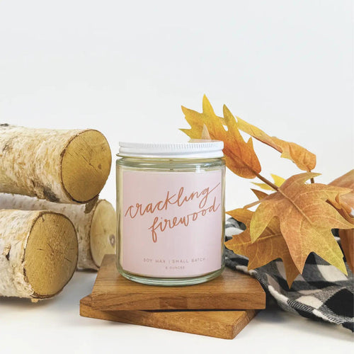 “Amber glass candle labeled CRACKLING FIREWOOD surrounded by pinecones and holiday décor.”

“Hand-poured soy candle burning beside a cozy fireplace and mug of cocoa.”

“Flat lay of CRACKLING FIREWOOD candle with matchsticks and festive greenery.”