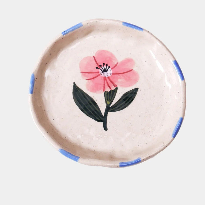 “Hand-painted stoneware plate with pink flower design and blue rim brushstrokes on a white background.”

“Close-up of small ceramic dish with pink floral design used to hold gold jewelry.”

“Delicate pink flower plate displayed on a pastel table with dried petals and a cup of tea.